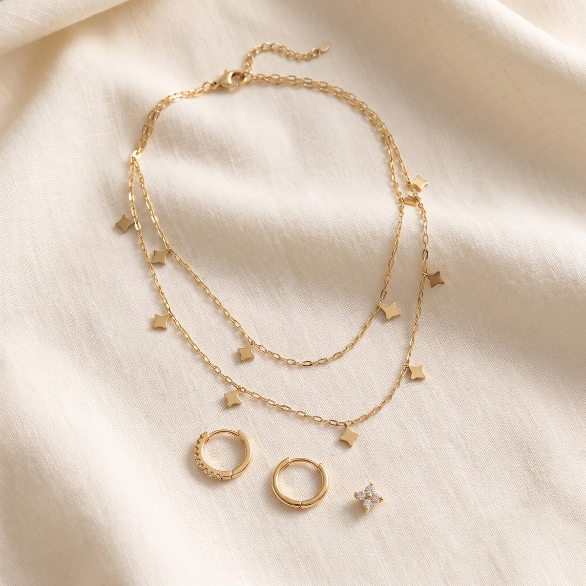 Layered gold tone necklace with star charms on ivory linen beside the Huggie earrings