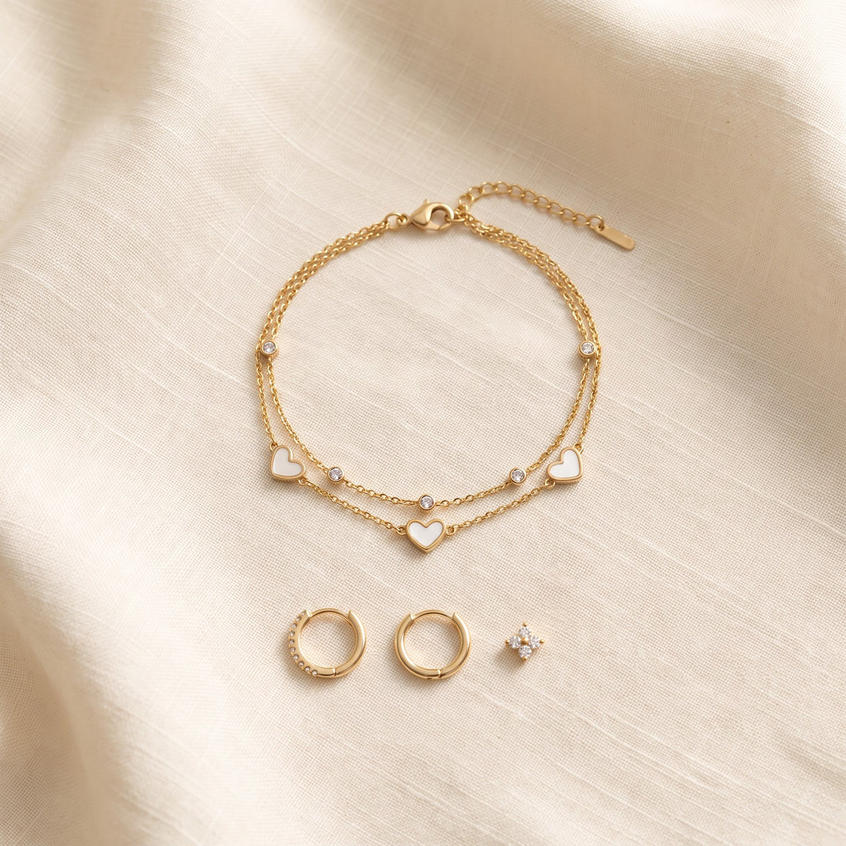 Gold tone heart bracelet on ivory linen beside the Huggie earrings
