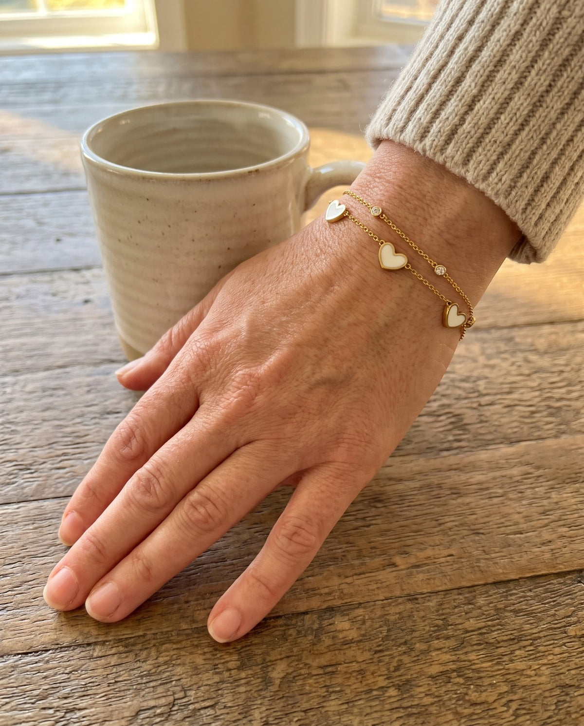 Gold tone heart bracelet worn on a wrist, a hand around a coffee mug